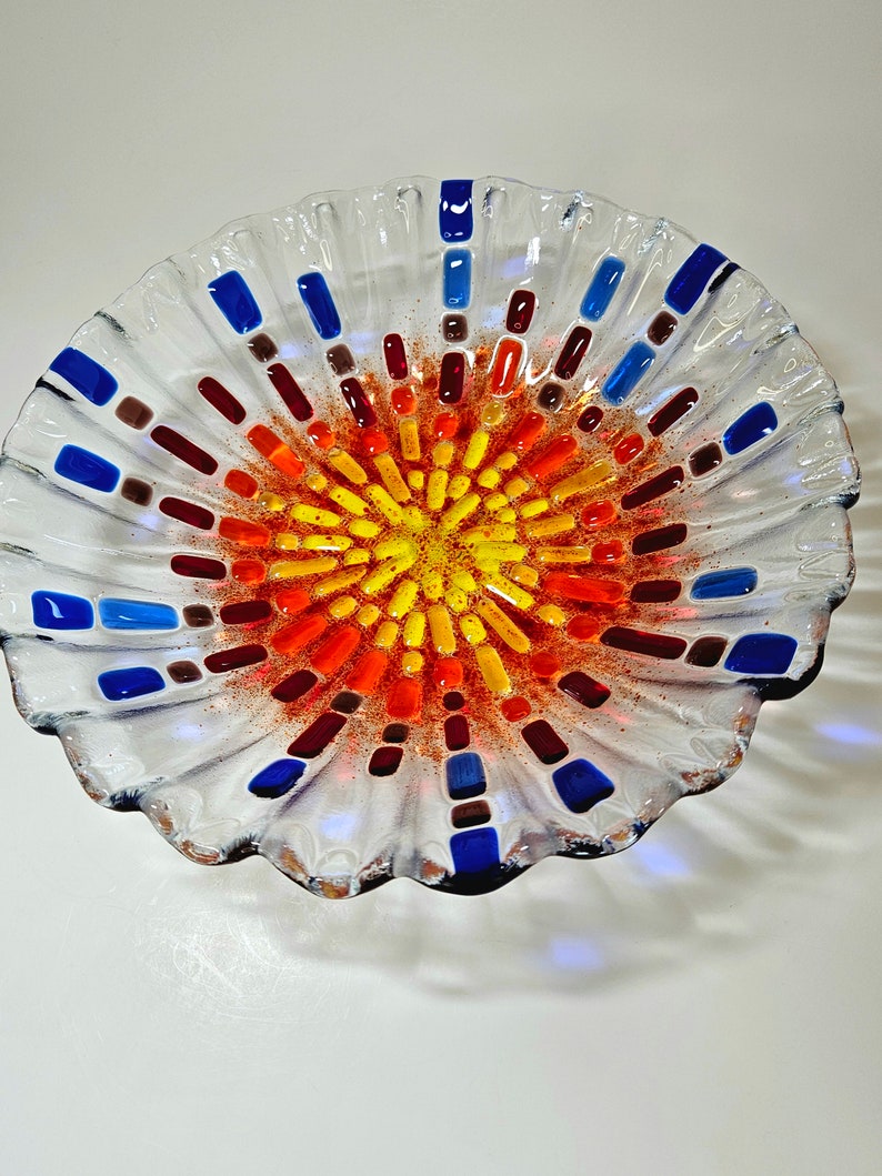 May include: A clear glass bowl with a scalloped edge and a colorful mosaic design in the center. The mosaic features shades of red, orange, yellow, blue, and brown.