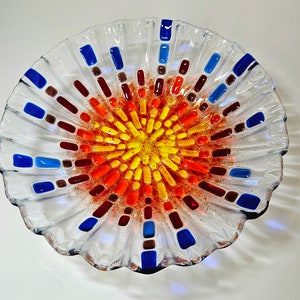 May include: A clear glass bowl with a scalloped edge and a colorful mosaic design in the center. The mosaic features shades of red, orange, yellow, and blue.