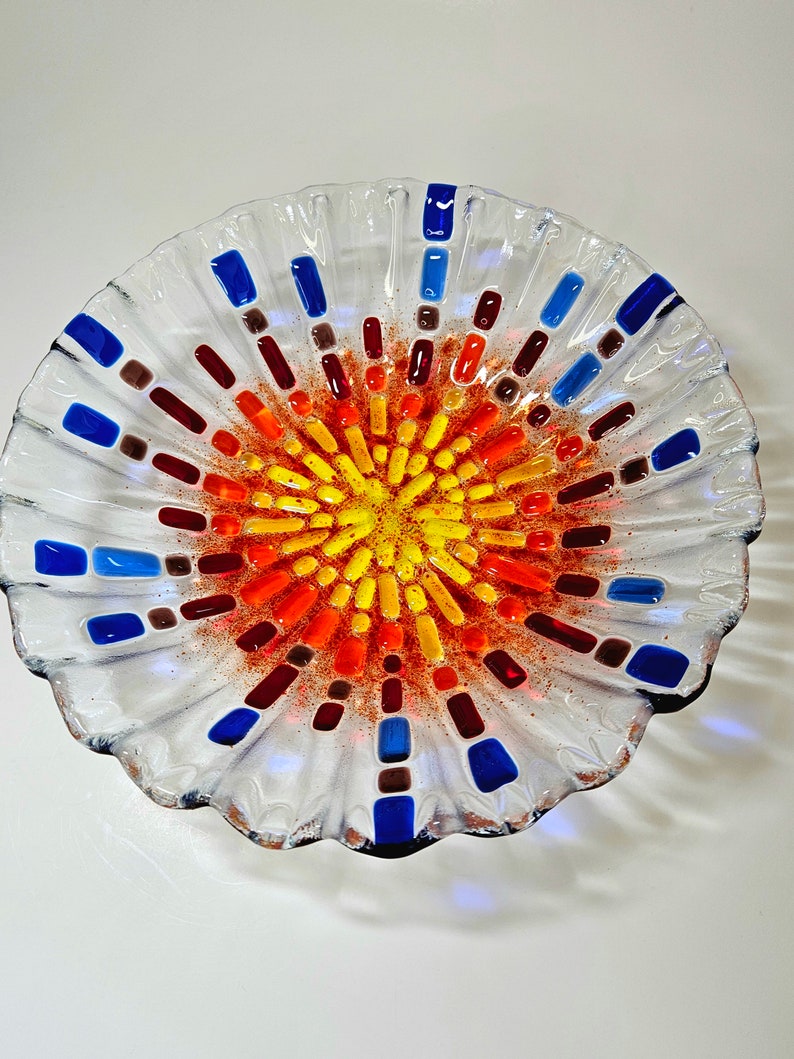 May include: A clear glass bowl with a scalloped edge and a colorful mosaic design in the center. The mosaic features shades of red, orange, yellow, and blue.