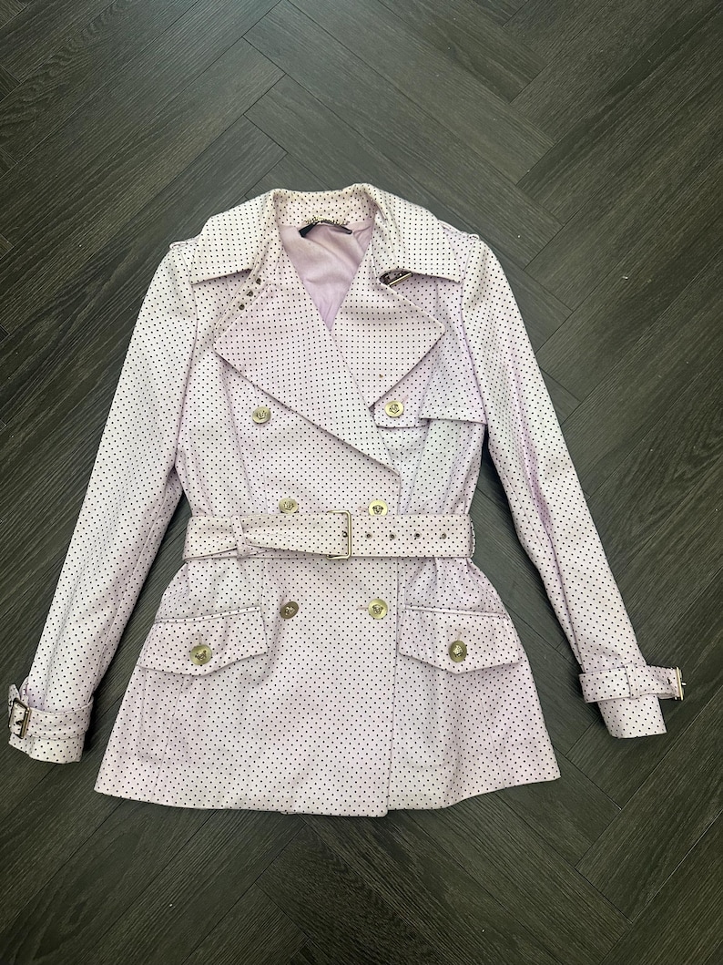 May include: A light purple trench coat with a polka dot pattern. The coat features a double-breasted design, a belt, and gold-colored buttons. The coat has two front pockets and a notched collar.