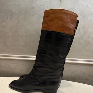 May include: Black and brown leather riding boots. The boots have a knee-high design with a brown leather upper and a black leather lower section. The boots have a low heel and a classic, equestrian-inspired style.
