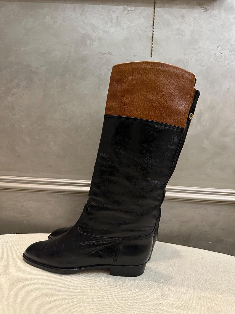 May include: Black and brown leather riding boots. The boots have a knee-high design with a brown leather upper and a black leather lower section. The boots have a low heel and a classic, equestrian-inspired style.