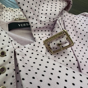 May include: Close-up of a light purple fabric with black polka dots. The fabric is part of a garment, featuring a collar and a metal buckle. The label reads "VERSA".