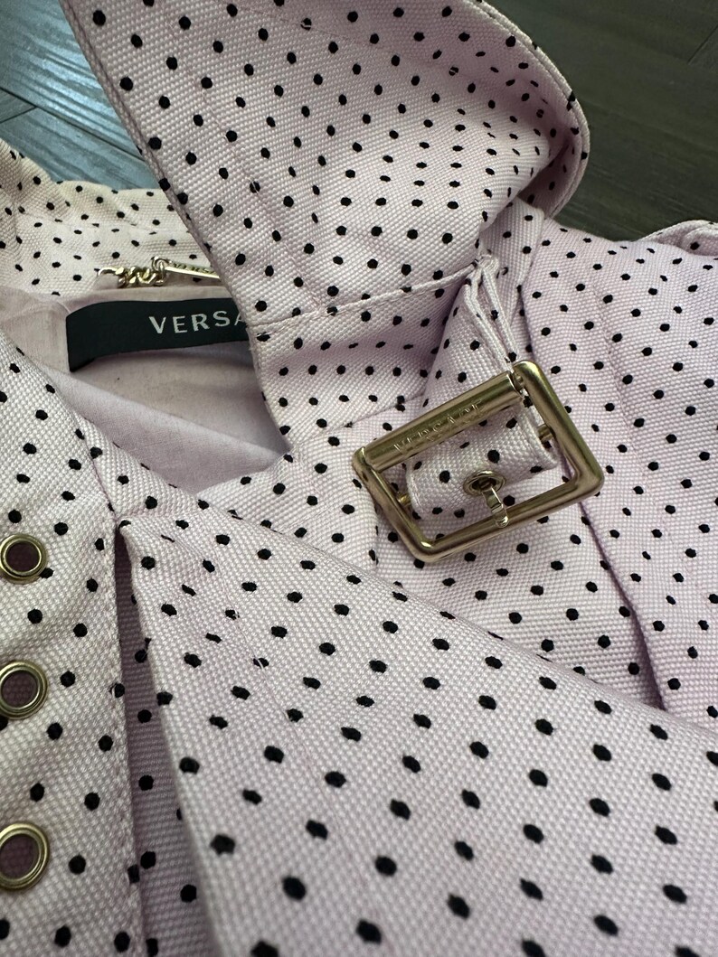 May include: Close-up of a light purple fabric with black polka dots. The fabric is part of a garment, featuring a collar and a metal buckle. The label reads "VERSA".