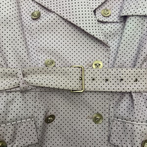 May include: Close-up of a light purple trench coat with a black polka dot pattern. The coat features a double-breasted design, a belt with a gold buckle, and gold buttons with a crest design. The coat is a stylish outerwear garment.