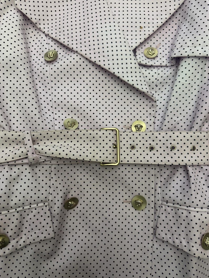 May include: Close-up of a light purple trench coat with a black polka dot pattern. The coat features a double-breasted design, a belt with a gold buckle, and gold buttons with a crest design. The coat is a stylish outerwear garment.