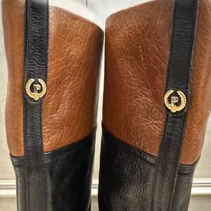 May include: A pair of knee-high boots with a two-tone design. The boots feature a black lower section and a brown upper section. A black strap with a gold emblem is on the side of each boot. The boots are made of leather.