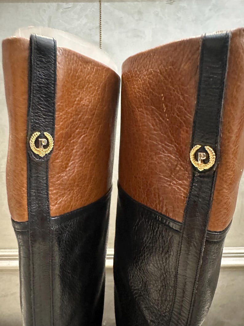 May include: A pair of knee-high boots with a two-tone design. The boots feature a black lower section and a brown upper section. A black strap with a gold emblem is on the side of each boot. The boots are made of leather.