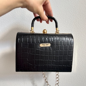 May include: Black rectangular handbag with a crocodile skin texture. The bag features a gold clasp, handle attachments, and a chain. The handle is black, and the bag has a small gold label that reads "Ruby Rose Collection."