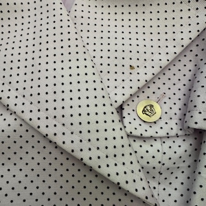 May include: Close-up of a light-colored blazer with a polka dot pattern. The blazer features a gold button with a detailed emblem. The fabric has a subtle texture, and the design is classic and elegant.