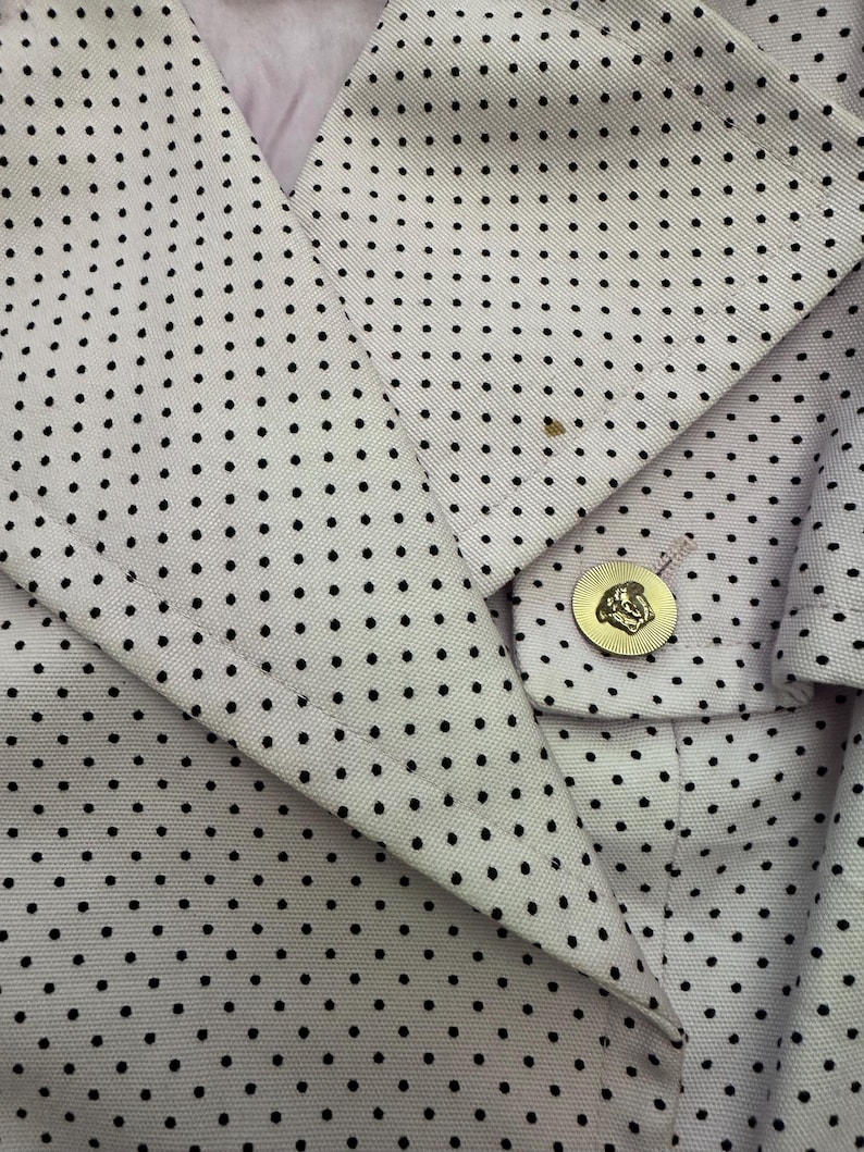 May include: Close-up of a light-colored blazer with a polka dot pattern. The blazer features a gold button with a detailed emblem. The fabric has a subtle texture, and the design is classic and elegant.