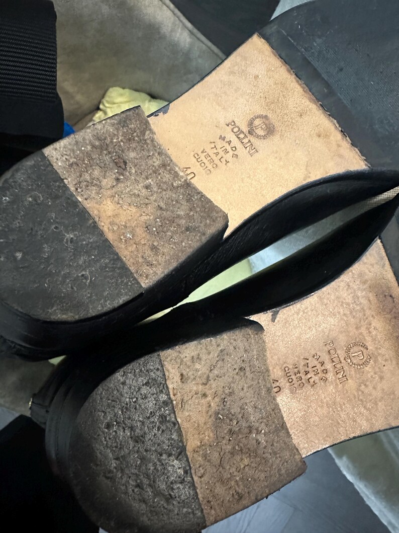 May include: Black leather shoes with worn soles. The soles are marked "POLLINI MADE IN ITALY VERO CUOIO 40". The shoes are likely Italian-made, with a focus on quality and craftsmanship.