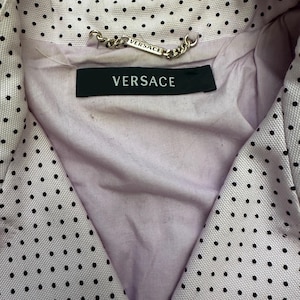 May include: Close-up of a light pink blazer with black polka dots. The blazer has a V-neck design and a black label with the word "VERSACE" in white. A silver chain with the word "VERSACE" is attached to the collar.