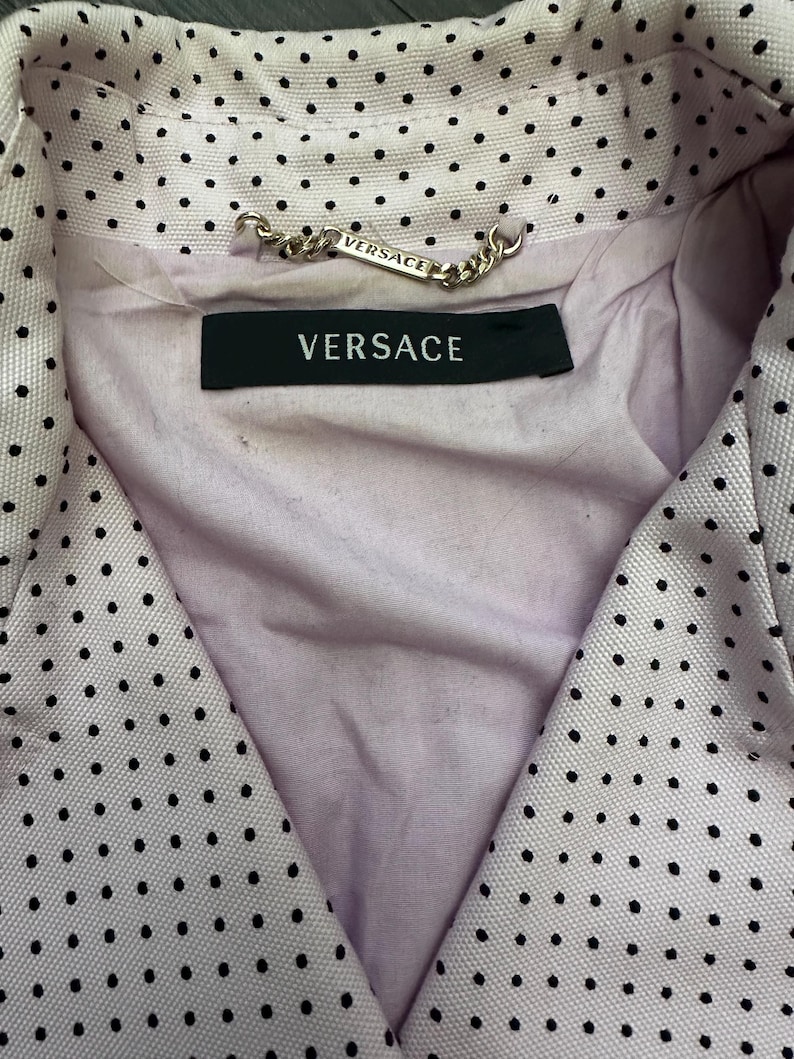 May include: Close-up of a light pink blazer with black polka dots. The blazer has a V-neck design and a black label with the word "VERSACE" in white. A silver chain with the word "VERSACE" is attached to the collar.