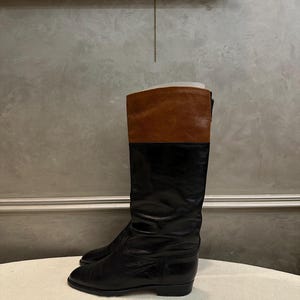 May include: A pair of black leather riding boots with a brown leather upper. The boots have a classic design, with a tall shaft and a low heel. The boots are displayed on a neutral surface.