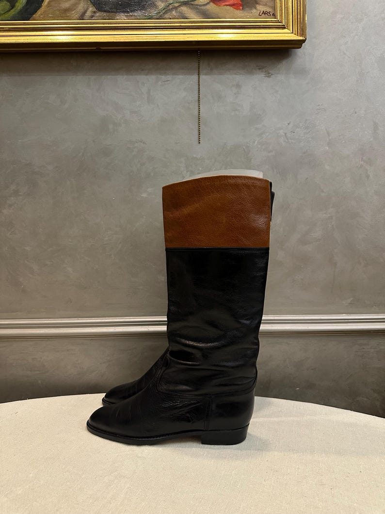 May include: A pair of black leather riding boots with a brown leather upper. The boots have a classic design, with a tall shaft and a low heel. The boots are displayed on a neutral surface.