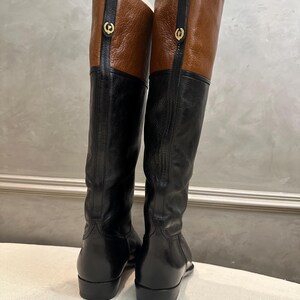 May include: A pair of tall black leather boots with brown leather tops. The boots have a gold-colored emblem on the side and are knee-high. The boots are displayed against a neutral background.