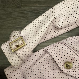May include: Close-up of a light pink jacket with black polka dots. The jacket features a gold-colored buckle on the sleeve and a gold button with a Medusa head emblem. The fabric has a textured appearance.