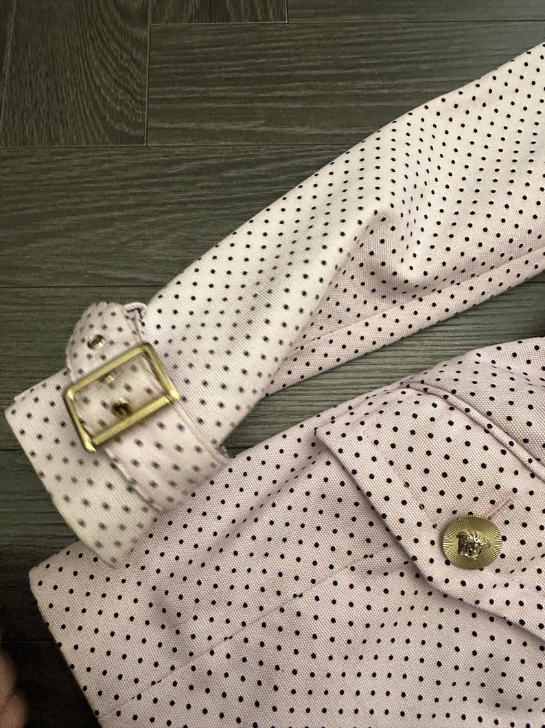 May include: Close-up of a light pink jacket with black polka dots. The jacket features a gold-colored buckle on the sleeve and a gold button with a Medusa head emblem. The fabric has a textured appearance.