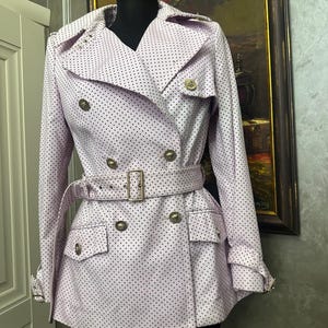 May include: A light pink trench coat with a polka dot pattern. The double-breasted coat features gold-colored buttons, a matching belt, and pockets. The coat is displayed on a black mannequin.