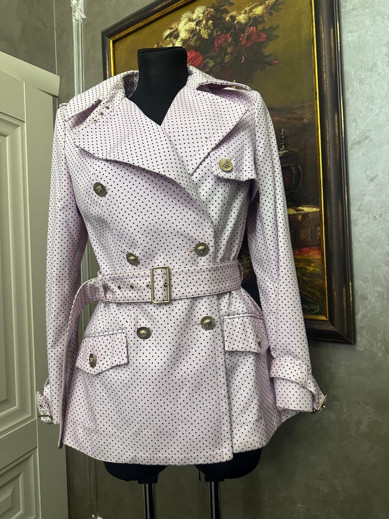 May include: A light pink trench coat with a polka dot pattern. The double-breasted coat features gold-colored buttons, a matching belt, and pockets. The coat is displayed on a black mannequin.
