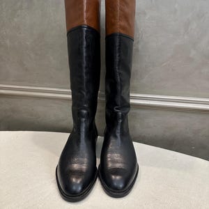May include: A pair of knee-high boots with a two-tone design. The boots feature a black lower section and a brown upper section. The boots are made of leather and have a classic, equestrian style.