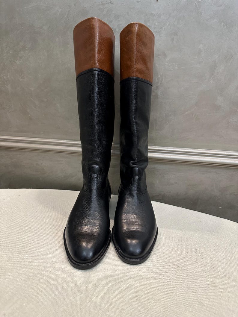 May include: A pair of knee-high boots with a two-tone design. The boots feature a black lower section and a brown upper section. The boots are made of leather and have a classic, equestrian style.