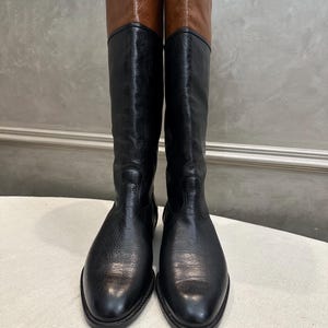 May include: A pair of tall, black leather boots with brown leather tops. The boots have a classic design, with a smooth, polished finish. The boots are displayed upright, showcasing their full height and shape. The boots are suitable for various occasions.