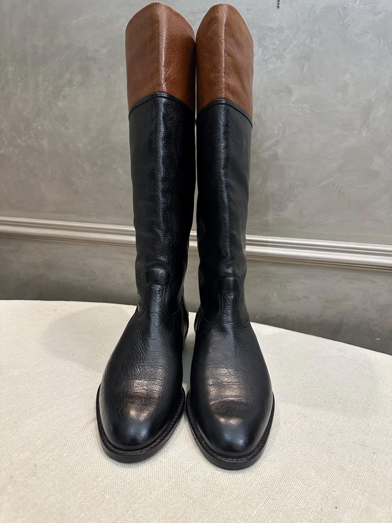 May include: A pair of tall, black leather boots with brown leather tops. The boots have a classic design, with a smooth, polished finish. The boots are displayed upright, showcasing their full height and shape. The boots are suitable for various occasions.