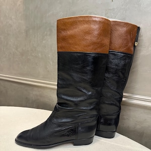 May include: A pair of knee-high boots with a color-block design. The boots feature a black lower section and a brown upper section. The boots have a low heel and a smooth leather finish. The boots are displayed on a white surface.