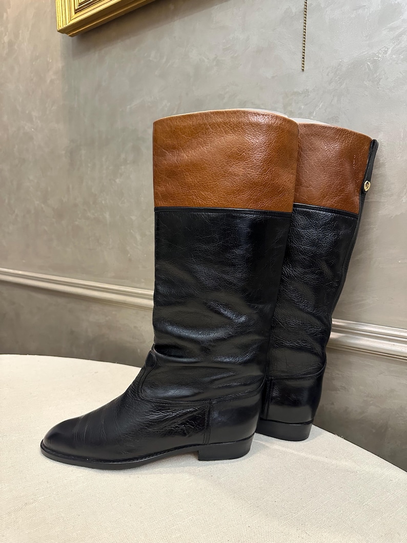 May include: A pair of knee-high boots with a color-block design. The boots feature a black lower section and a brown upper section. The boots have a low heel and a smooth leather finish. The boots are displayed on a white surface.