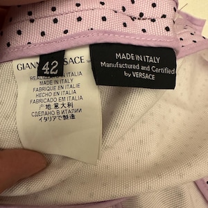 May include: Close-up of a garment's interior, featuring labels with text in multiple languages, including "MADE IN ITALY" and "Manufactured and Certified by VERSACE." The garment has a light purple lining with black polka dots.