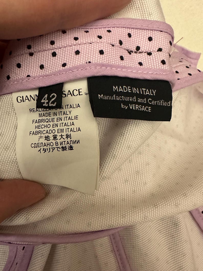 May include: Close-up of a garment's interior, featuring labels with text in multiple languages, including "MADE IN ITALY" and "Manufactured and Certified by VERSACE." The garment has a light purple lining with black polka dots.