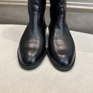 May include: A pair of black leather boots. The boots have a classic design with a rounded toe and a mid-calf height. The leather has a textured appearance, and the boots appear to be unworn.