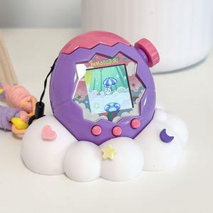 May include: A purple Tamagotchi virtual pet with a pink top and a cloud-shaped base. The screen displays a cartoon character. The base has a pink heart, a yellow star, and a blue crescent moon. A colorful lanyard is attached.