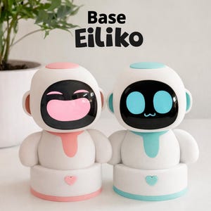 Op de afbeelding: Twee witte robotspeelgoed met zwarte schermen en kleurrijke accenten. Eén heeft een roze gezicht en accenten, de andere blauw. De tekst "Base Eiliko" staat erboven. Elke robot heeft een hart op zijn basis.
