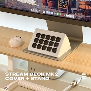 May include: A Stream Deck MK2 cover and stand. The control panel is cream-coloured with sixteen dark square buttons. The stand is silver, and a small, white, pig-shaped figurine sits to the left. The text "STREAM DECK MK2 COVER + STAND" is at the bottom.