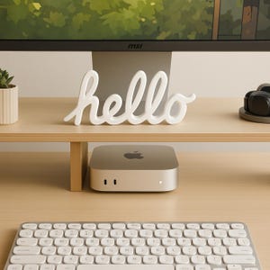 May include: A white 'hello' decorative sign sits on a wooden shelf above a silver computer. A keyboard is in the foreground. A small plant in a white pot is on the left. The computer has the Apple logo.