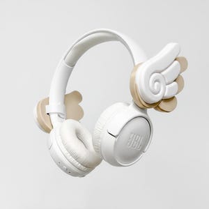 Può includere: Cuffie JBL bianche con dettagli a forma di ali beige. Le cuffie hanno una fascia e padiglioni imbottiti. Il logo JBL è visibile sui padiglioni. I dettagli alati sono sui lati.