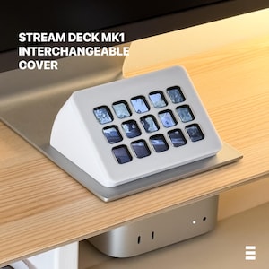 Op de afbeelding: Een witte Stream Deck MK1 verwisselbare cover met 15 vierkante knoppen, elk met een kleine afbeelding. Het apparaat staat op een zilveren basis, met de tekst "STREAM DECK MK1 INTERCHANGEABLE COVER" erboven.