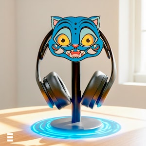 Könnte beinhalten: Ein schwarzer Kopfhörerständer mit einem blauen Katzen-Gesicht-Design. Der Ständer hält schwarze Kopfhörer und hat eine kreisförmige Basis mit blauem Licht. Das Katzengesicht ist blau mit gelben Augen und weißen Zähnen.