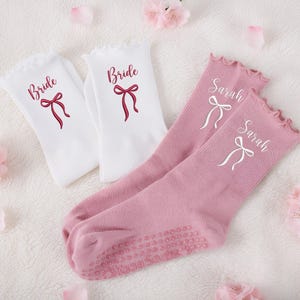 Könnte beinhalten: Weiße und rosa Socken mit Rüschen am oberen Rand. Die weißen Socken haben das Wort "Bride" und ein rotes Schleifendesign. Die rosa Socken sind mit dem Namen "Sarah" und einem weißen Schleifendesign personalisiert. Hochzeitsthema.