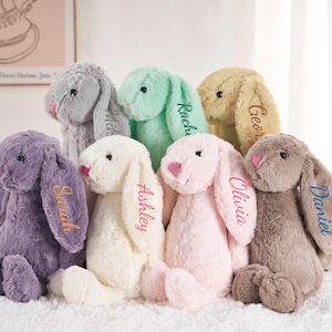 May include: A collection of plush bunny rabbits in various pastel colors, each with a name embroidered on one ear. The bunnies have pink noses and are arranged together. The names include Sarah, Ashley, Olivia, and Daniel.