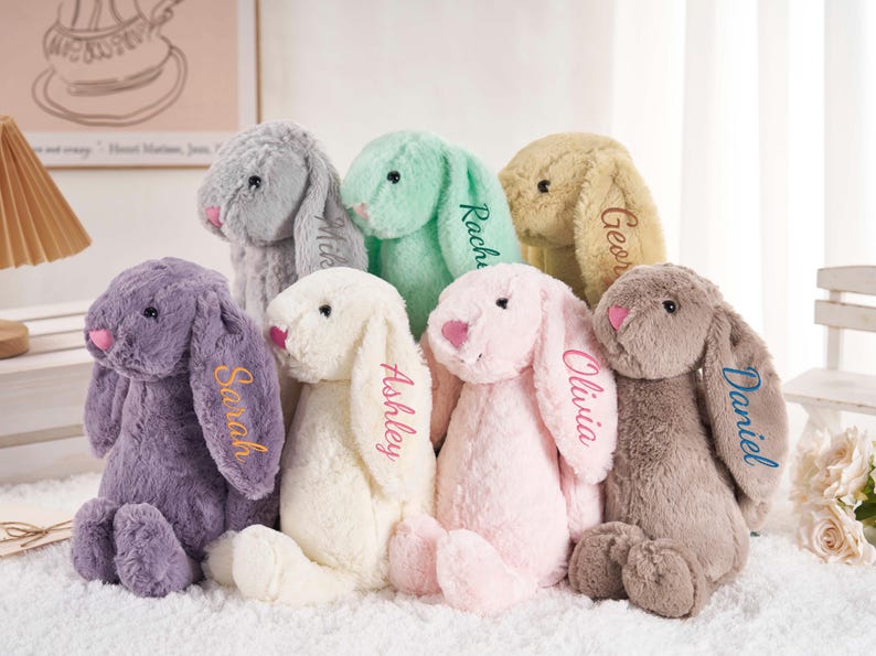 May include: A collection of plush bunny rabbits in various pastel colors, each with a name embroidered on one ear. The bunnies have pink noses and are arranged together. The names include Sarah, Ashley, Olivia, and Daniel.