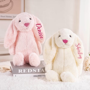 May include: Two plush bunny rabbits, one pink and one cream-colored, sit side-by-side. The pink bunny has "Denise" embroidered on its ear, while the cream bunny has "Shirley." Both have pink noses and are on a white surface.