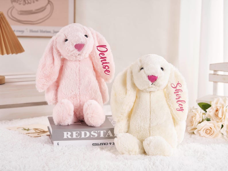 May include: Two plush bunny rabbits, one pink and one cream-colored, sit side-by-side. The pink bunny has "Denise" embroidered on its ear, while the cream bunny has "Shirley." Both have pink noses and are on a white surface.