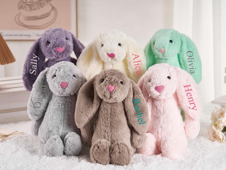 May include: A collection of six plush bunny rabbits in various pastel colors: lavender, cream, mint green, gray, brown, and pink. Each bunny has a pink nose and the name of a person embroidered on one ear. The bunnies are soft and cuddly.
