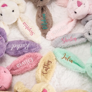 May include: A collection of plush bunny rabbits in various pastel colors, including pink, purple, and mint green. Each bunny has an ear embroidered with a name, such as "Sophia" or "Julia". The bunnies are arranged on a soft, white surface.