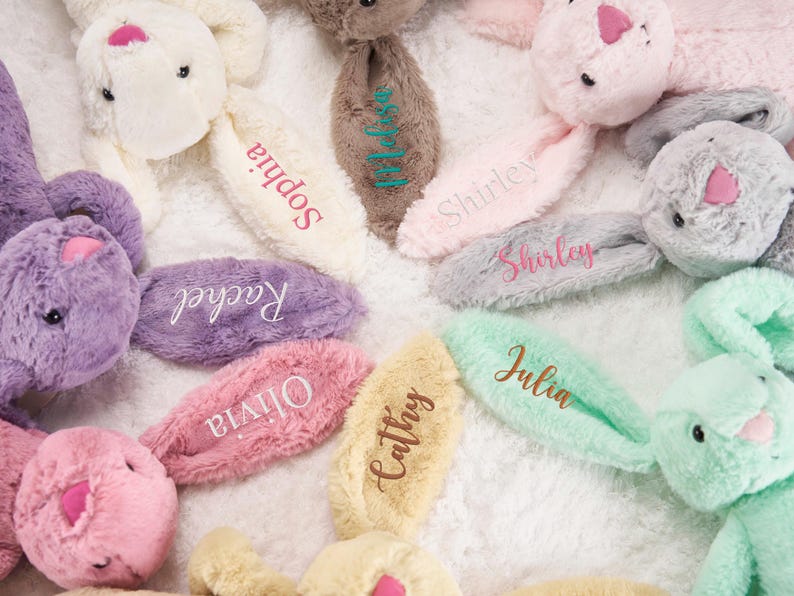 May include: A collection of plush bunny rabbits in various pastel colors, including pink, purple, and mint green. Each bunny has an ear embroidered with a name, such as "Sophia" or "Julia". The bunnies are arranged on a soft, white surface.
