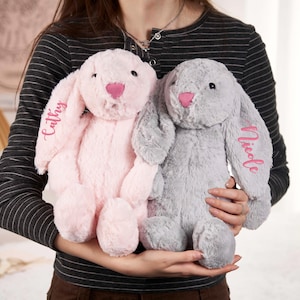 May include: Two plush bunny rabbits, one pink and one gray, are held in front of a person. The pink bunny has the name "Cathy" embroidered on its ear, and the gray bunny has "Nicole" embroidered on its ear. Both bunnies have pink noses.